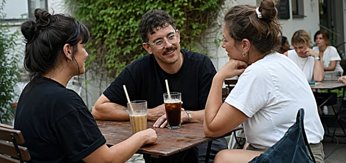 Drei Gaeste an einem Holztisch auf einer Cafe-Terrasse, Cold Brew und Iced Latte, Pflanzenwand im Hintergrund, sommerliche Treffpunkt-Atmosphaere